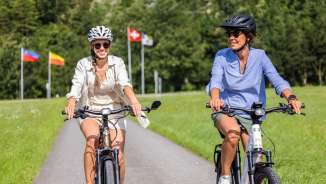 Two women on e-bikes on a summer tour with the flags of Liechtenstein, Switzerland and Austria in the background